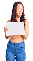 Fototapeta premium Young beautiful chinese girl holding paper banner with blank space serious face thinking about question with hand on chin, thoughtful about confusing idea