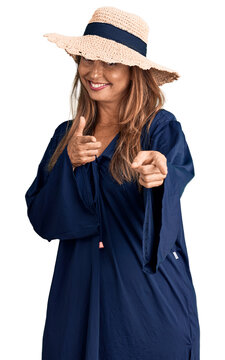 Middle age hispanic woman wearing summer hat pointing fingers to camera with happy and funny face. good energy and vibes.