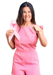 Young beautiful brunette woman holding pink cancer ribbon annoyed and frustrated shouting with...