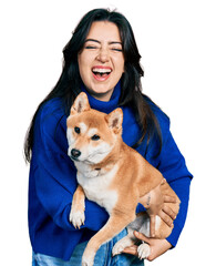 Beautiful hispanic woman holding cute dog smiling and laughing hard out loud because funny crazy...