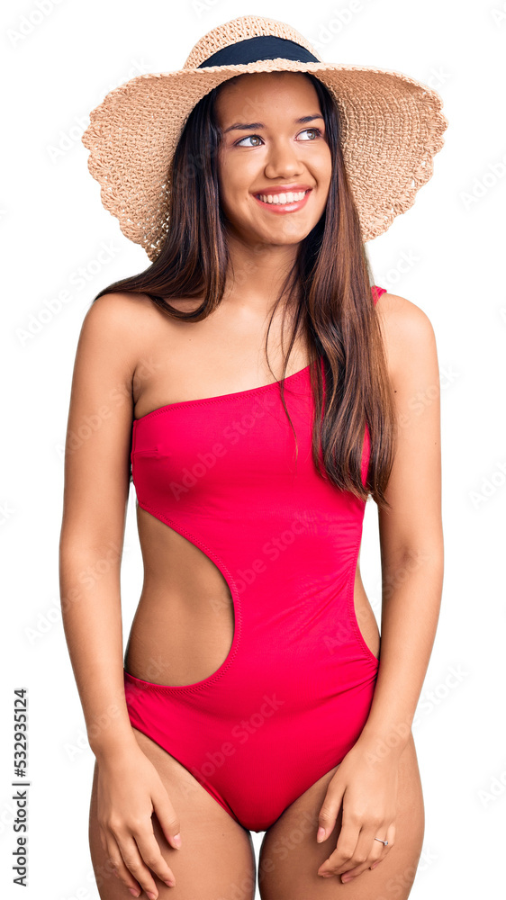 Wall mural young beautiful latin girl wearing swimwear and summer hat looking away to side with smile on face, 