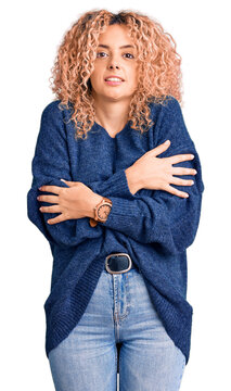 Young blonde woman with curly hair wearing casual winter sweater shaking and freezing for winter cold with sad and shock expression on face