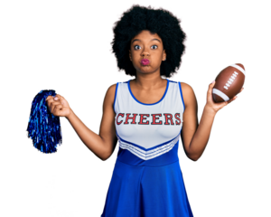 Young african american woman wearing cheerleader uniform holding pompom and football ball puffing cheeks with funny face. mouth inflated with air, catching air.