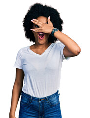 Young african american woman wearing casual white t shirt peeking in shock covering face and eyes with hand, looking through fingers with embarrassed expression.