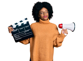 Young african american woman holding video film clapboard and megaphone smiling looking to the side...