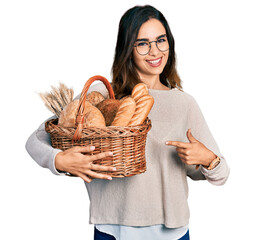Beautiful hispanic woman holding wicker basket with bread smiling happy pointing with hand and...