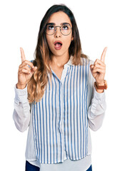 Beautiful hispanic woman wearing casual striped shirt amazed and surprised looking up and pointing with fingers and raised arms.