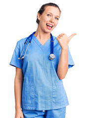 Young beautiful blonde woman wearing doctor uniform and stethoscope smiling with happy face looking and pointing to the side with thumb up.