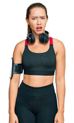 Beautiful blonde woman wearing gym clothes and using headphones in shock face, looking skeptical and sarcastic, surprised with open mouth
