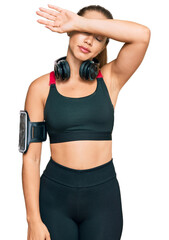 Beautiful blonde woman wearing gym clothes and using headphones covering eyes with arm, looking serious and sad. sightless, hiding and rejection concept