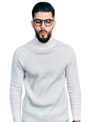 Young arab man with beard wearing elegant turtleneck sweater and glasses making fish face with...