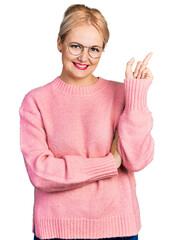 Young blonde woman wearing casual clothes and glasses with a big smile on face, pointing with hand and finger to the side looking at the camera.