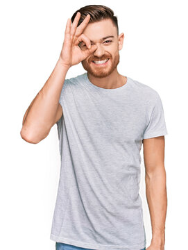 Young redhead man wearing casual grey t shirt doing ok gesture with hand smiling, eye looking through fingers with happy face.
