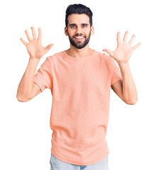 Young handsome man with beard wearing casual t-shirt showing and pointing up with fingers number ten while smiling confident and happy.