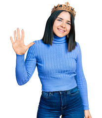 Young brunette woman with blue eyes wearing princess crown showing and pointing up with fingers number five while smiling confident and happy.