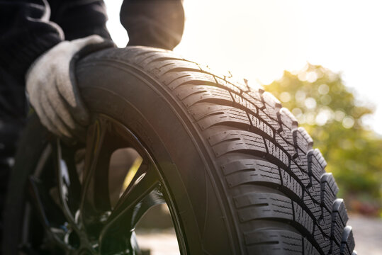Close-up of hands with gloves rolling a new winter tyre  in bright sunlight ready for tyre change. Driving safety, road safety and car service concept.