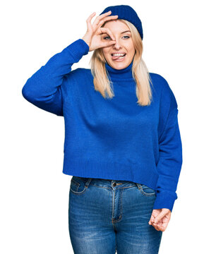 Young caucasian woman wearing wool winter sweater and cap doing ok gesture like binoculars sticking tongue out, eyes looking through fingers. crazy expression.