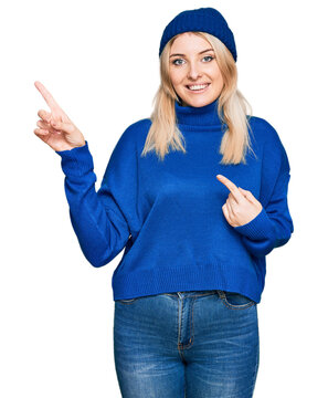Young caucasian woman wearing wool winter sweater and cap smiling and looking at the camera pointing with two hands and fingers to the side.
