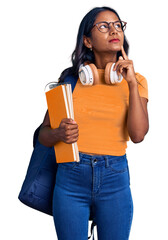 Young indian girl holding student backpack and books serious face thinking about question with hand...