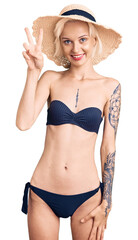 Young blonde woman with tattoo wearing bikini and summer hat showing and pointing up with fingers number two while smiling confident and happy.