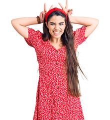 Brunette teenager girl wearing summer dress posing funny and crazy with fingers on head as bunny...