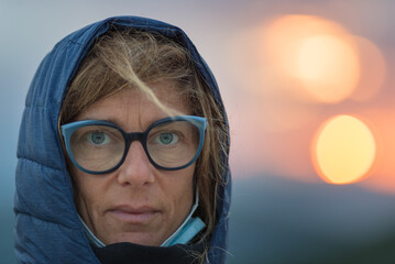 Outdoors portrait of adult mid age woman with blue green eyes, wearing glasses. Defocused backgropund, serious facial expression.