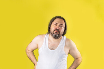 A disgruntled, unshaven man in a white undershirt stands with his hands on his hips and looks ahead incredulously. Unemployed slovenly dressed man from Eastern Europe.