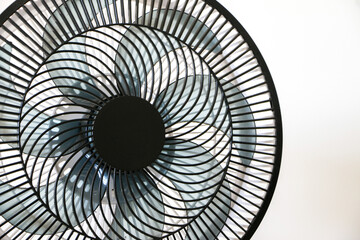 Close up of rotating black electric fan showing its spinning blades, on left of frame, against white wall, necessary appliance for summer heat waves and rising temperatures