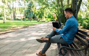 Obraz premium Serious young adult businessman, sitting in the park, turning on his laptop.