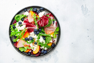 Fresh salad with arugula, radicchio, peaches, smoked pork ham, feta cheese and blueberries. White table background, top view.