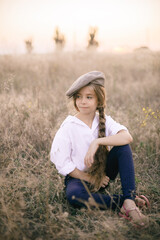 
cute little girl with a long braid of blond hair in a white shirt and a men's vintage cap at sunset in a summer field.