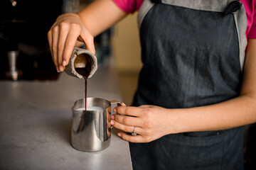 Fototapeta na wymiar Female hand carefully pours coffee from grey measuring cup into metal jug