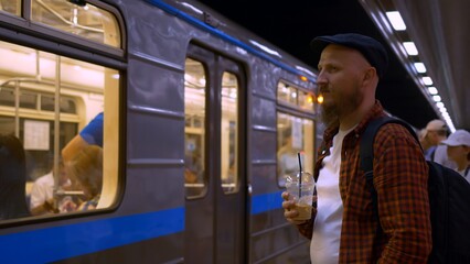 A man with a beard in a cap is waiting for a subway train, drinking cold coffee with ice. A wagon pulls up to the underground subway platform. The headlights of a subway train illuminate a man.