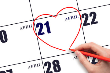 A woman's hand drawing a red heart shape on the calendar date of 21 April. Heart as a symbol of...