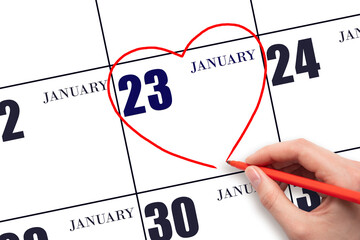 A woman's hand drawing a red heart shape on the calendar date of 23 January. Heart as a symbol of...