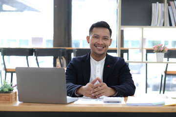 Fototapeta na wymiar Portrait of a young Asian businessman smiling while using a laptop and writing down notes while sitting at his desk in a modern office
