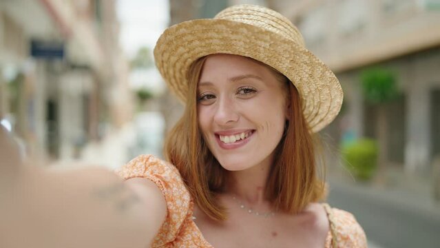 Young redhead woman tourist wearing summer hat make selfie by the camera at street