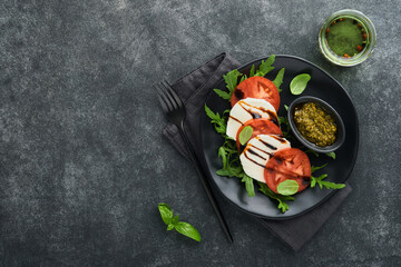 Caprese salad. Italian caprese salad with sliced tomatoes, mozzarella cheese, arugula, basil, olive oil in black plate over old brick tiles black background. Delicious Italian food. Top view.