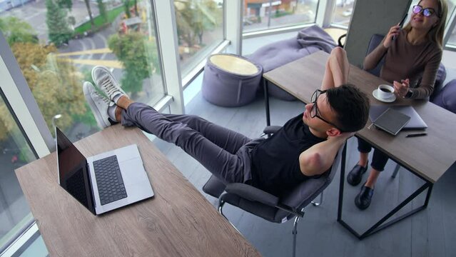People having a break from work in the office. Man sits with his legs on the table and hands behind his head. Woman next table speaking on the phone. High angle view. City in window.