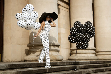 Spring, summer fashion. Glamour, stylish elegant woman in polka dot jumpsuit and big hat is holding balloons with dots. Fashion model in outfit with polka dots in the city. 60's style. Retro fashion