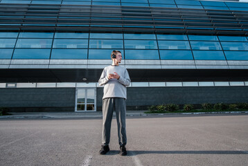 Young stylish man using the phone outdoors. Successful young teenager walking outside office building,  smiling, using headphones for music with  phone.