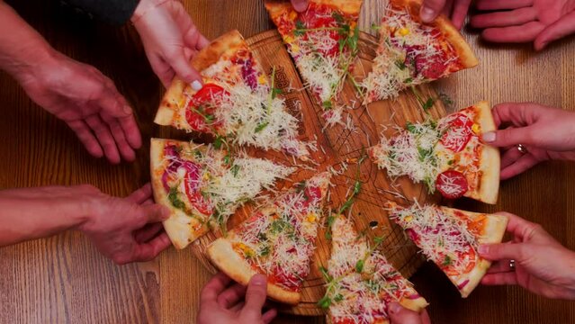 Top view male and female hands taking slices of pizza with cheese, tomatoes and ham from food delivery. Group of hungry friends sitting at desk and sharing delicious lunch on wooden table background