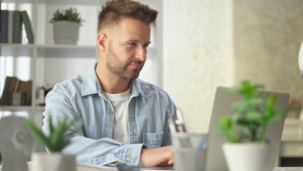 Happy businessman in casual using laptop in home office, Young adult man sitting at desk in study room, working online with computer, browsing the Internet, smiling.