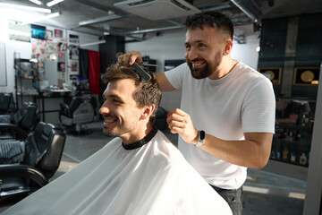 Obraz premium Cheerful barbershop worker smiling while combing young brunet guy hair