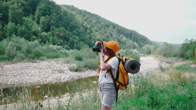 A young woman takes pictures on a DSLR camera. Hiking with camera, orange backpack and cap. A tourist girl takes pictures of nature during a trip. A trip to rest in nature in the forest and mountains.