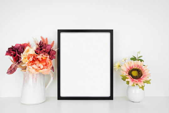 Mock up black picture frame with pink hue autumn flowers on a wood shelf against a white wall. Fall concept. Copy space.