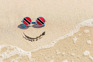 Fototapeta na wymiar A painted smile on the beach and sunglasses with the flag of Trinidad and Tobago. The concept of a positive holiday in the resort of Trinidad and Tobago.