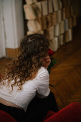 Fototapeta premium Woman with curly hair sits from back and listens against the background of books, holding rose