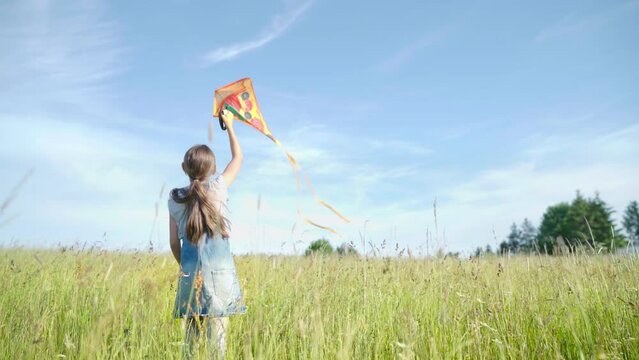 4K little girl flying bright orange kite in bright blue sky pulling on rope. Childhood, spending time outdoor, leisure activities, games concept.