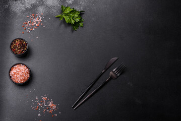 An empty black plate on a dark concrete background with spices, herbs and cutlery